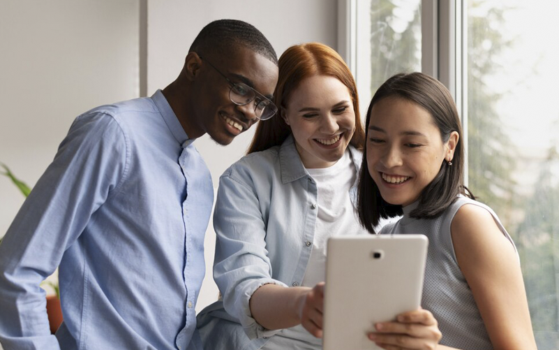 Imagem de capa de blog post sobre recrutamento da geração Z nas empresas. Na foto, há 3 jovens trabalhando. Eles estão sorrindo enquanto analisam informações em um tablet.