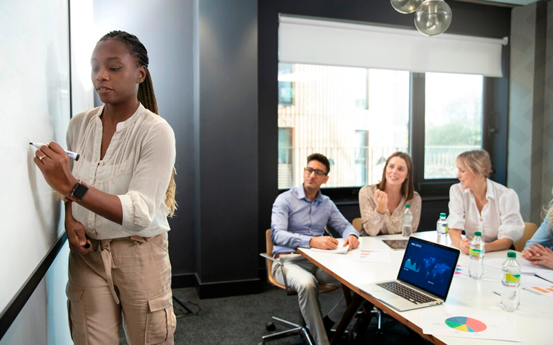 Imagem de capa de blog post sobre liderança e cultura nas empresas. Na foto, há a simulação de um treinamento, onde uma mulher é a líder do grupo e ensina para outras três pessoas. Ela está a frente da sala e escreve em um quadro.