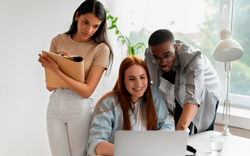 Imagem de capa de blog post sobre o perfil da geração z no trabalho. Na foto, 3 jovens (duas mulheres e um homem) conversam sobre uma ideia apresentada em um notebook.